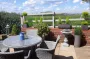 A stone terrace in the sunshine with a view over green fields and a table and chair set in the foreground.